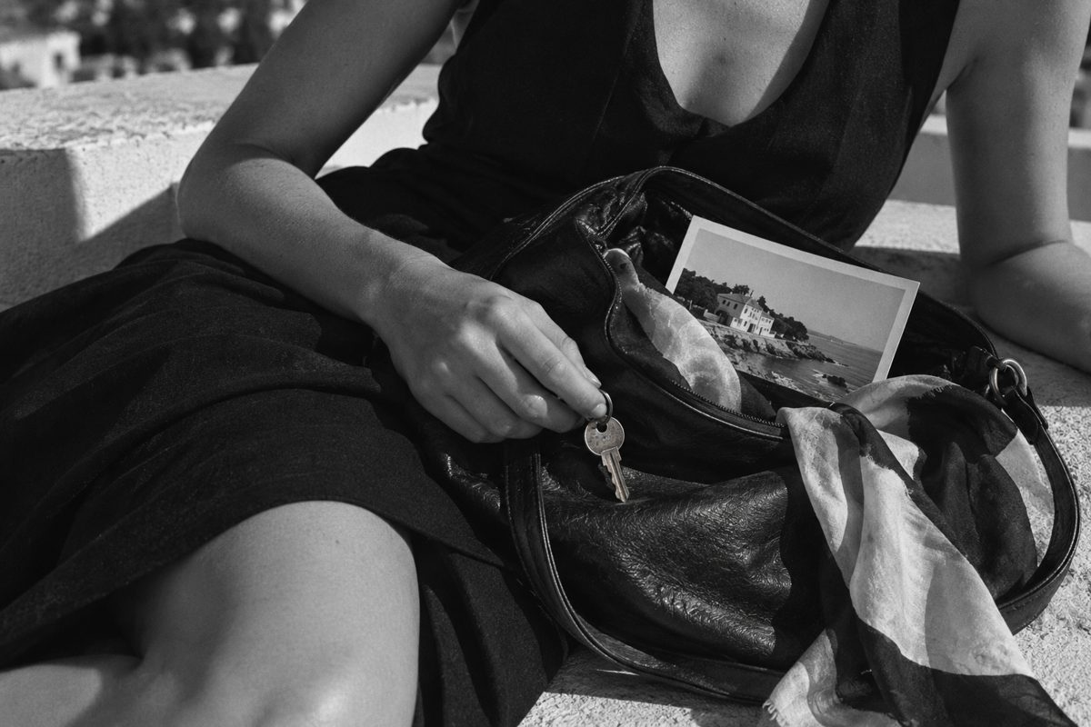 A hand holding a house key over a leather bag containing a coastal photograph and silk scarf.