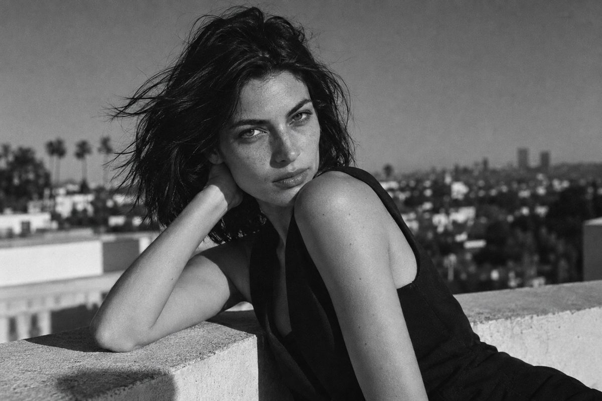 A woman with windblown dark hair in a black dress leaning against a sunlit terrace wall.